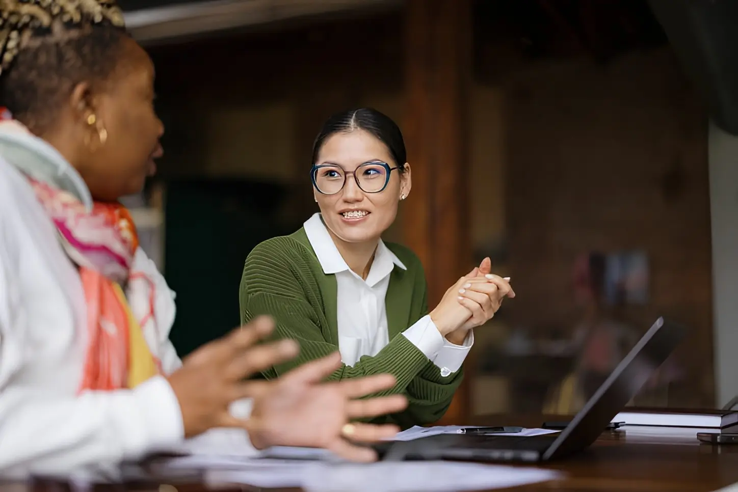 Two women engaged in conversation at a table, with a laptop open between them.